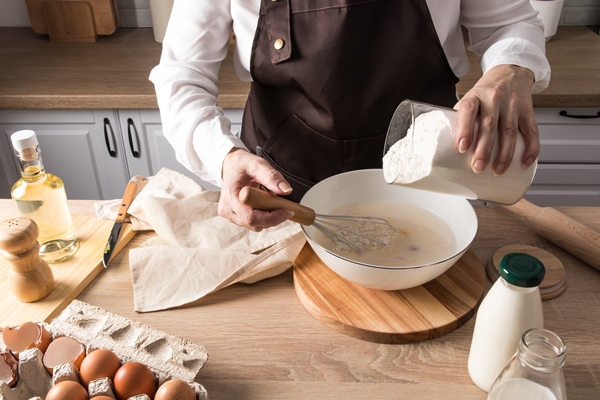 the hands of a woman in an apron pour flour from a jar into a bowl of milk the concept of cooking dough at home in the kitchen - Заливной пирог с зелёным луком и яйцом