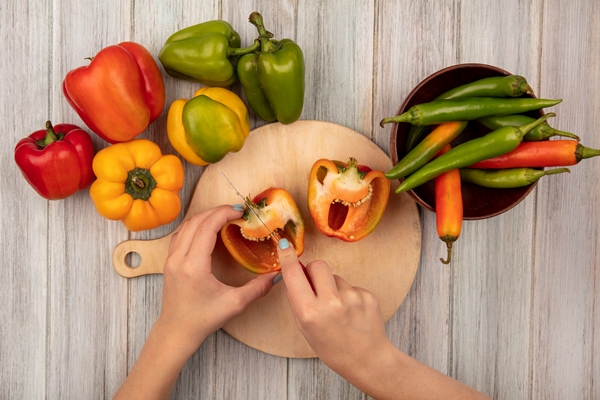 top view of female hands cutting orange bell peppers on a wooden kitchen board with knife on a grey wooden surface - Аджика с яблоками