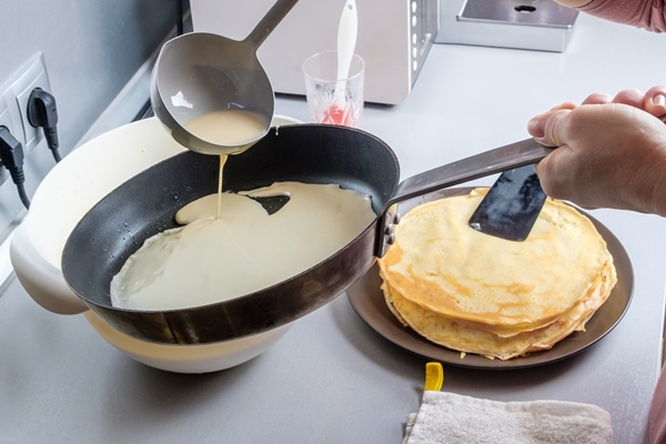 batter pouring out of a metal bucket onto a red hot greased frying pan the process of baking pancakes traditional treat for the holiday of maslenitsa - Постные блинчики на рисовом отваре с овощами