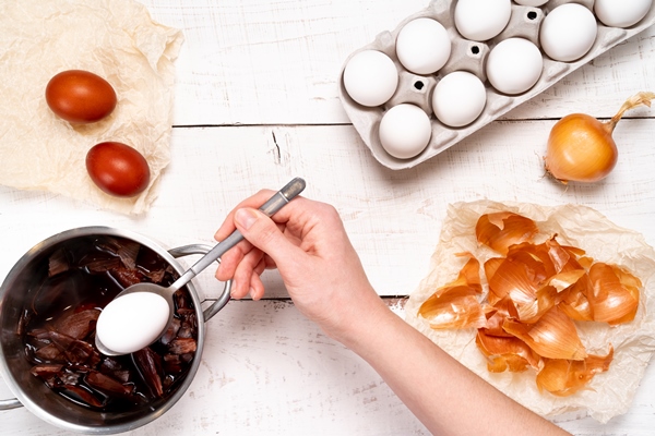 easter eggs the process of coloring with natural dye onion husks in a small saucepan on a white wooden table and a woman s hand holding a egg in a spoon - Яйца, крашенные луковой шелухой