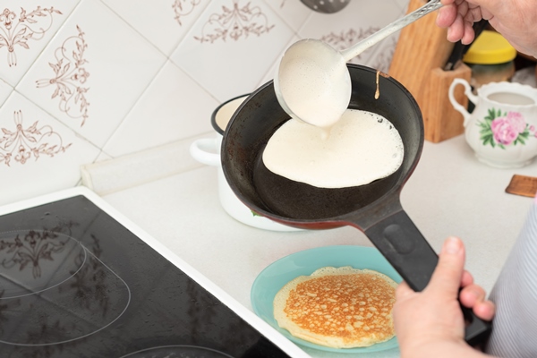 senior woman chef pour portion of liquid dough by hands with ladle on frying pan for baking pancakes - Постные гречневые блины на закваске