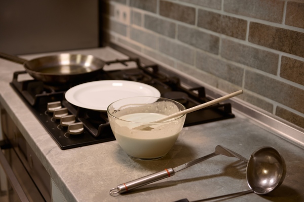 wooden spoon in glass bowl with pancake batter stainless steel kitchen utensils on kitchen countertop white plate and frying pan on a black stove - Постные ванильные блинчики