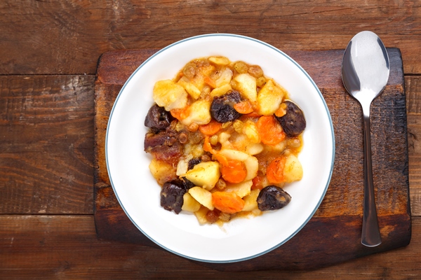 a dish of jewish cuisine tsimes with carrots and dates in a white plate on a wooden board next to a spoon - Тушёный картофель с черносливом и изюмом