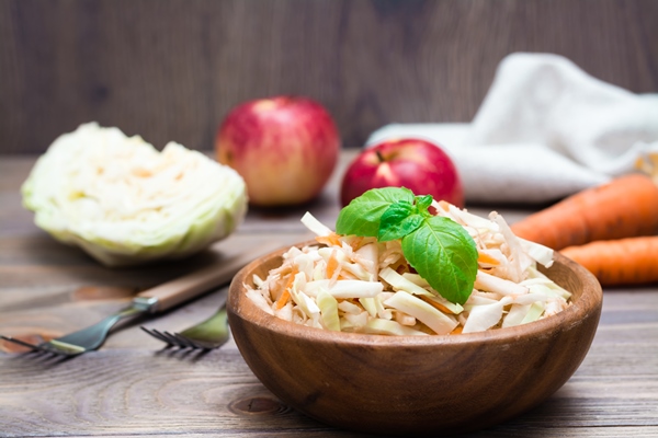 american ready to eat cole slaw salad made from cabbage celery carrots and apples with basil leaves in a wooden plate and ingredients for cooking on a wooden table - Салат из капусты с яблоками