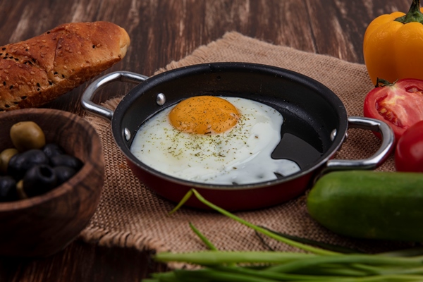 side view fried eggs in a frying pan with green onions olives tomatoes cucumbers and a loaf of bread on a wooden background - Бутерброд с яйцом, помидором и зеленью