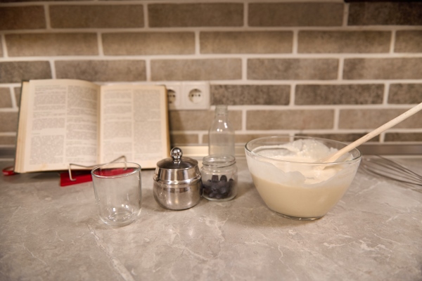 transparent glass bowl with wooden spoon dropped into a liquid pancake dough blueberries in a glass container on the background of a recipe book on a kitchen countertop - Русские постные блины гречнево-пшеничные