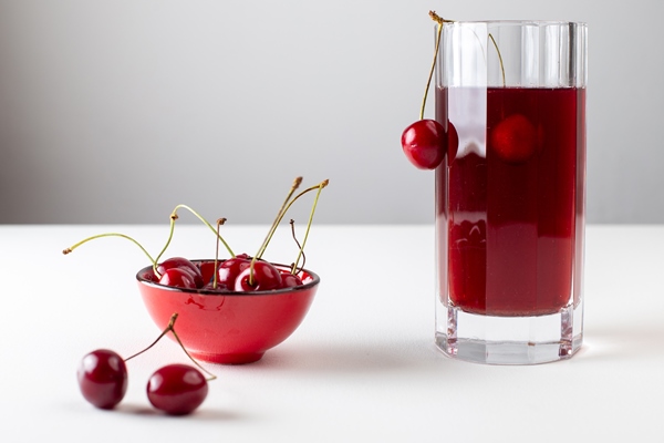 front view of cherry juice inside long glass with fresh cherries on the white surface - Вишнёвый квас