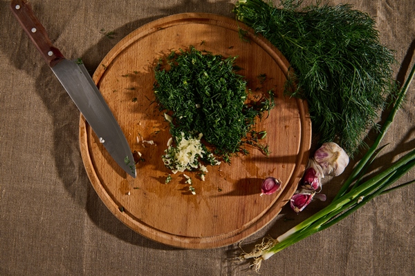 overhead view of a bunch of fresh moist dill on a linen tablecloth and garlic next to a wooden cutting board with a kitchen knife and chopped garlic and dill seasonal greens food background flat lay - Щи красные