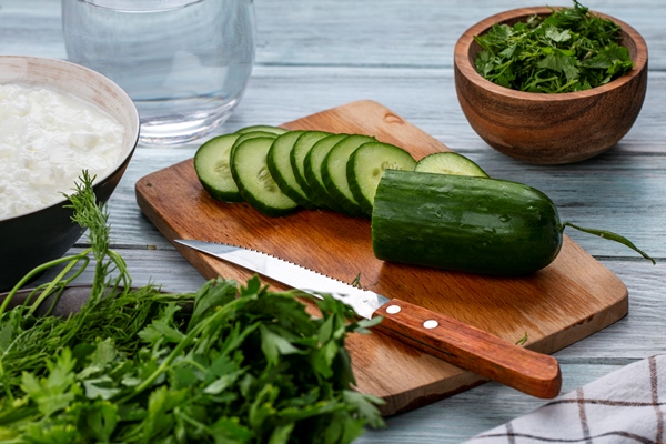 side view of sliced cucumbers on a board with a knife and greens on a gray surface - Постная окрошка