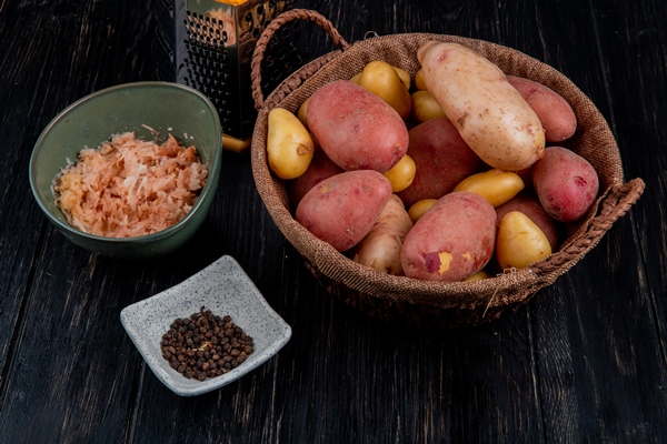 side view of whole potatoes in basket and grated ones in bowl with black pepper seeds and grater on wooden table - Драники с морковью и луком
