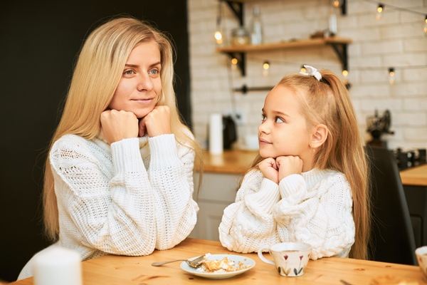 attractive young female with blonde long hair and her beautiful daughter both in cozy sweaters having breakfast in kitchen sitting at dining table drinking tea having cake keeping hands under chin - Учим детей заваривать чай