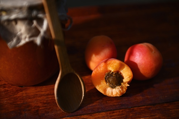 focus on halves of sweet ripe red sunny apricot with pit next to wooden tea spoon and jam jar on a rustic wooden surface - Сливовое варенье с косточками