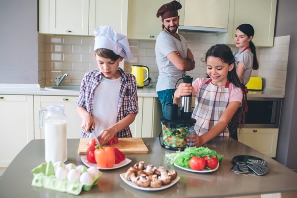 independent kids are preparing salad together without parents girl is mixing vegetables in blender while boy is cutting pieces of tomatoes parents look at them and controll - Грибы жареные, постный стол