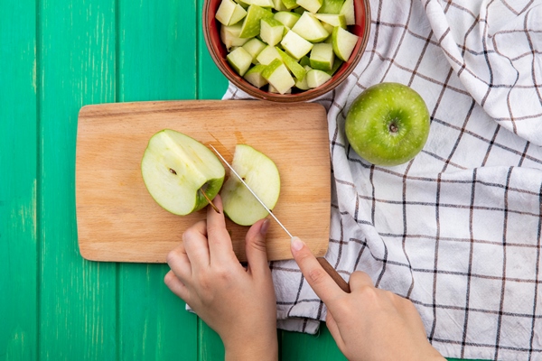 top view of female hands slicing green apple on wooden kitchen board on red bowl of chopped apples and cloth - Постная яблочная острая аджика