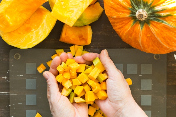 cutted pieces of pumpkin in woman hands on glass cutting board on wooden background - Постные оладьи из тыквы и банана