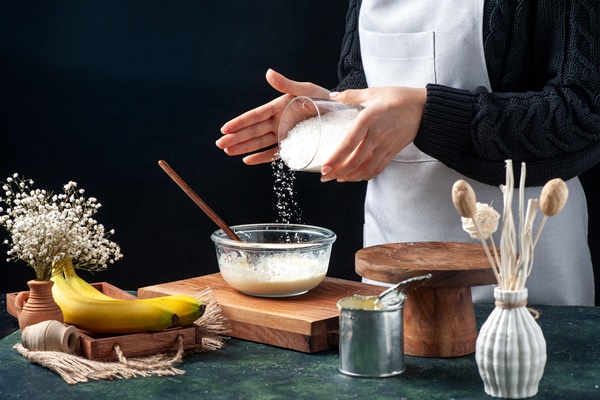 front view female cook pouring coconut powder onto condensed milk on a dark background - Постное кокосовое печенье