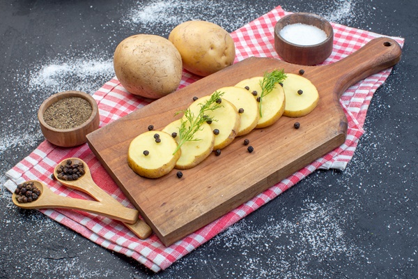 side view of fresh chopped potatoes with dill pepper on wooden cutting board on red stripped towel spices on dark color background - Сёмга, запечённая с луком и картофелем, постный стол