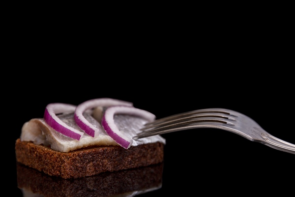 slices of herring on black bread with red onion rings and a fork lying next to it traditional russian appetizing appetizer for vodka close up black background with reflection space for - Бутерброды с малосольной сельдью и луком