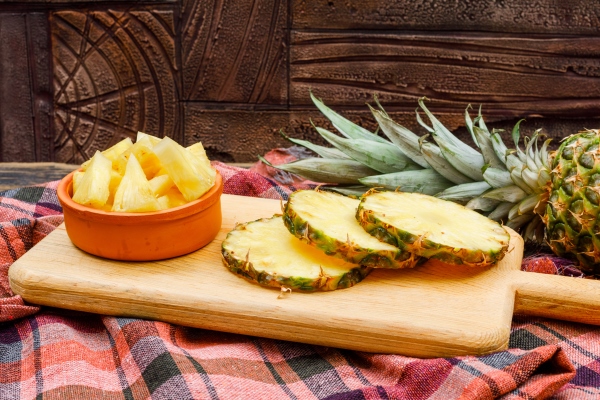 whole and sliced pineapple in a cutting board and clay bowl on a stone tile and picnic cloth side view - Пикантная закуска из ананаса