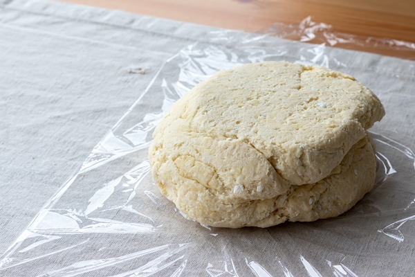 raw curd dough with bits of cottage cheese on a plastic wrap on a linen tablecloth on a wooden table - Ленивые вареники