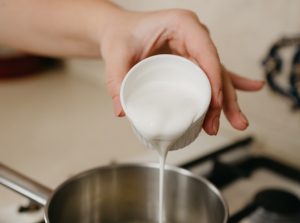 a photo of a hand of a woman who is pouring cream from the cup to the saucepan with lemon juice and zest on the gas stove - Сливочный суп с грибами и булгуром
