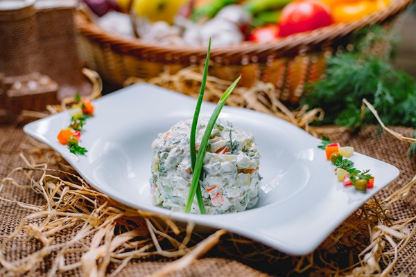 side view of traditional russian olivier salad with chicken decorated with green onion in a white bowl on straw background - Правила приготовления салатов