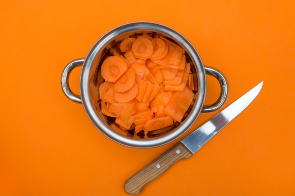 sliced carrots in a metal saucepan with a knife on an orange background - Творожники с морковью