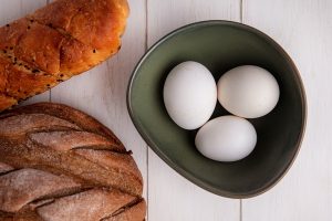 top view chicken eggs in bowl and loaf of black and white bread on white background - Паровое рыбное суфле
