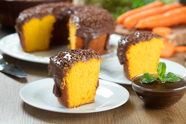 close up piece of brazilian carrot cake with chocolate frosting on wooden table with carrots in the background - Морковная бабка