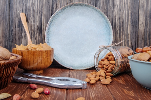 side view of almond scattered from a glass jar and a bowl with peanut butter on wooden background - Кокосово-миндальный урбеч