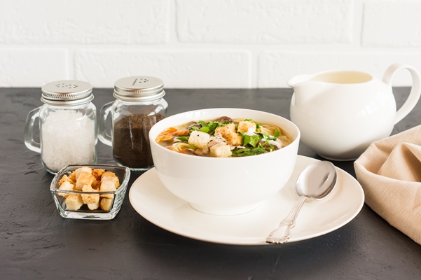 white mushroom soup with parsley cream and croutons in a soup plate on a black background opposite a white brick wall - Монастырская кухня: грибной бульон с расстегаями, кулеш