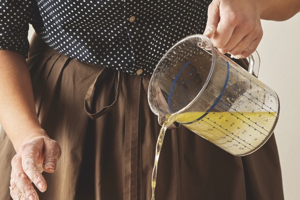 woman pours water with olive oil from measure cup to flour on board to prepare dough for pasta or dumplings cooking guide presentation - Монастырская кухня: вареники с яблоком и изюмом, морковное печенье