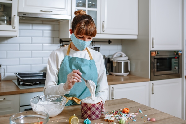 a cook in a medical mask and gloves in a home kitchen puts sweet figures on the icing of an easter cake - Куличи александрийские