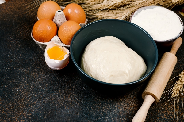 raw yeast dough in bowl and ingredients eggs flour ears of wheat on dark table bakery concept - Пасхальная мона