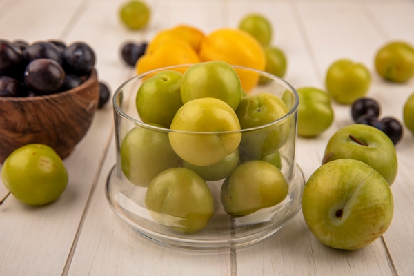 side view of fresh green cherry plums on a glass bowl with dark purple sloes on a wooden bowl with peaches isolated on a white wooden background - Повидло из лопуха без сахара
