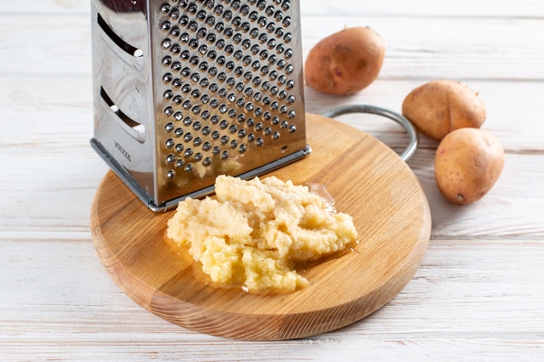 grated potato with metal standing grater on wooden cutting board on wooden table - Драники из картофеля и баклажанов