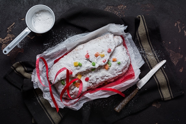 wholegrain stollen with raisins and powdered sugar on a linen napkin with a sieve red ribbon over the old dark concrete background traditional german christmas cake top view 1 - Масляный рождественский штоллен