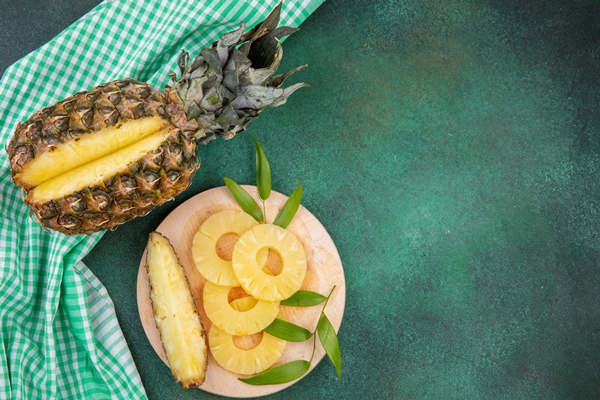 top view of pineapple with one piece cut out from whole fruit and pineapple slices on cutting board on plaid cloth and green surface - Смузи-боул с зелёным чаем