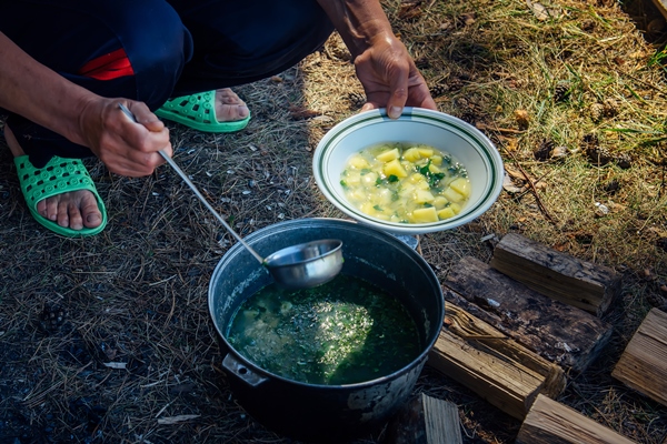 hands of adult woman pouring soup from a cauldron into a plate with ladle cooking in the fresh air on a hiking trip large pot standing on the grass and firewood concept of tourism - Организация трапезы в походе: хранение продуктов, походная кухня, утварь, меню