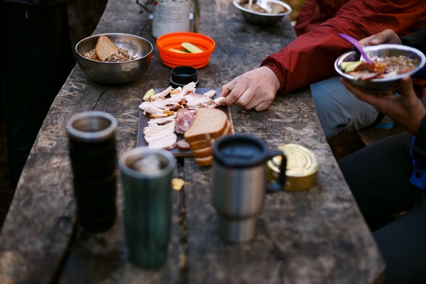 hiker s breakfast wooden table with bread bacon cans other meals and hot mugs at the forest camp - Питание в походе