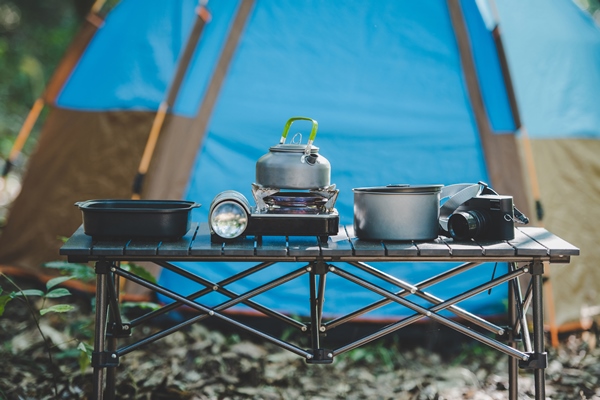 set of cooking equipment for camping trip with kettle pot pan flashlight mini gas stove and camera on table at front of camping tent in nature forest - Организация трапезы в походе: хранение продуктов, походная кухня, утварь, меню