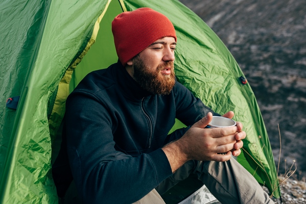 young explorer man drinking hot beverage in mountains sitting near to bonfire relaxing after trekking traveler man in red hat holding a mug of tea after hiking travel people - Питание в походе