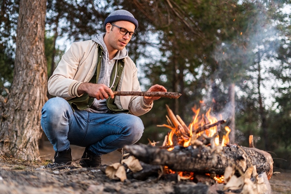 i want to keep warm handsome caucasian man in warm clothes making fire with sticks while squatting near dry branches - Пшеничная каша в котелке на костре