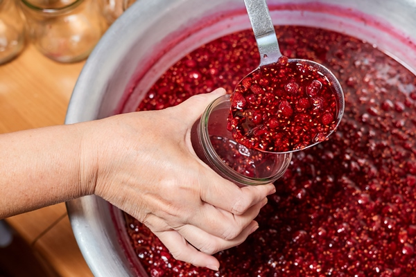 woman pouring freshly brewed raspberry jam into a glass jar using kitchen spoon - Малиновое варенье
