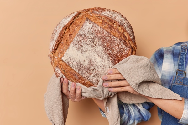 unrecognizable woman baker holds round fragrant rye bread on linen napkin demonstrates homemade freshly baked product against beige studio background rustic atmosphere rural bakery concept - Ржаной кислый хлеб