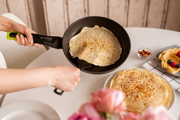 hands of young woman holding frying pan over kitchen table while taking hot appetizing pancake to put it on top of other crepes on plate - Блинцы-скородумки