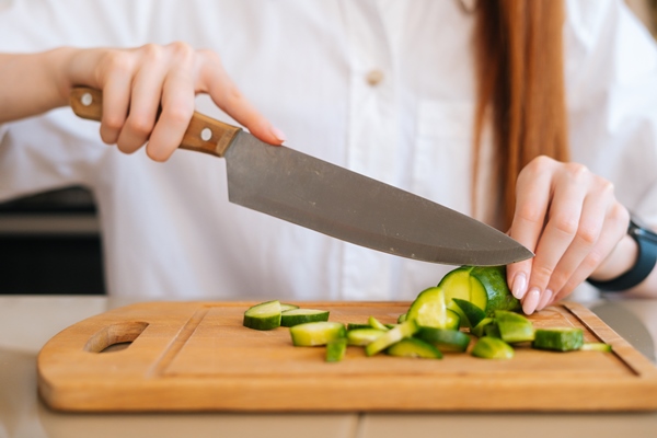 closeup front view of unrecognizable woman cutting fresh cucumber cooking food salad sitting at wodden table in light kitchen room front view of female cooking vegetarian dieting salad at home - Хозяйке на заметку: виды кухонных ножей